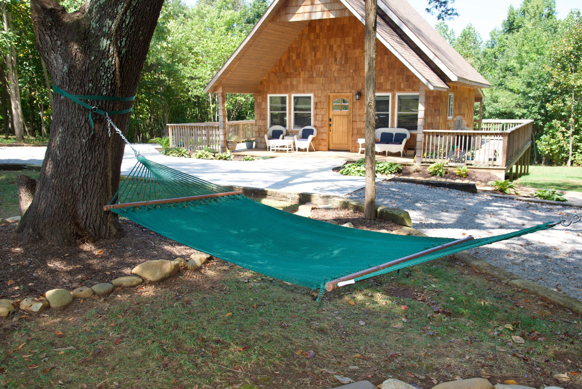 Green Double Caribbean Hammock hung between trees in front of a cedar-shingled cabin