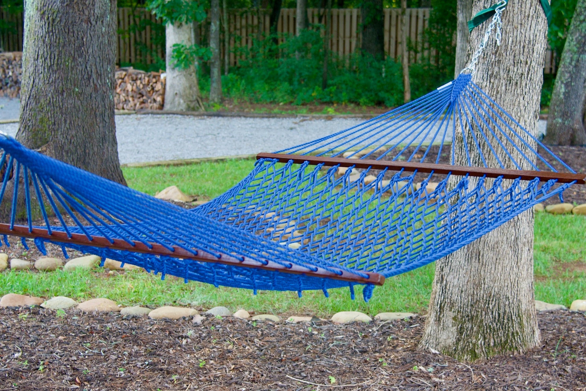 Dark blue Island Escape hammock hung between two trees above a stone-edged mulch bed, firewood stacked behind