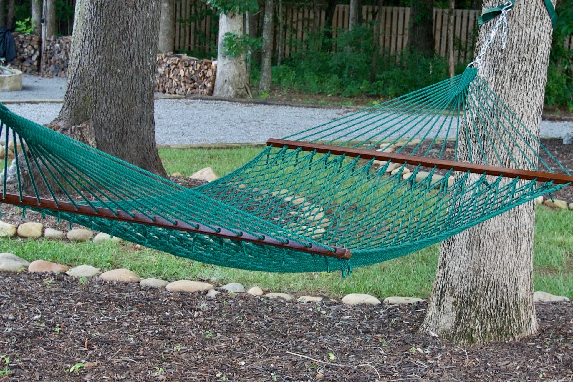 Green Island Escape hammock hung between two trees above a stone-edged mulch bed, firewood stacked behind
