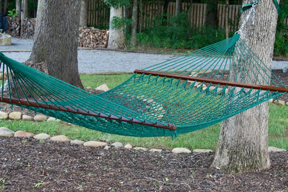 Green Island Escape hammock hung between two trees above a stone-edged mulch bed, firewood stacked behind