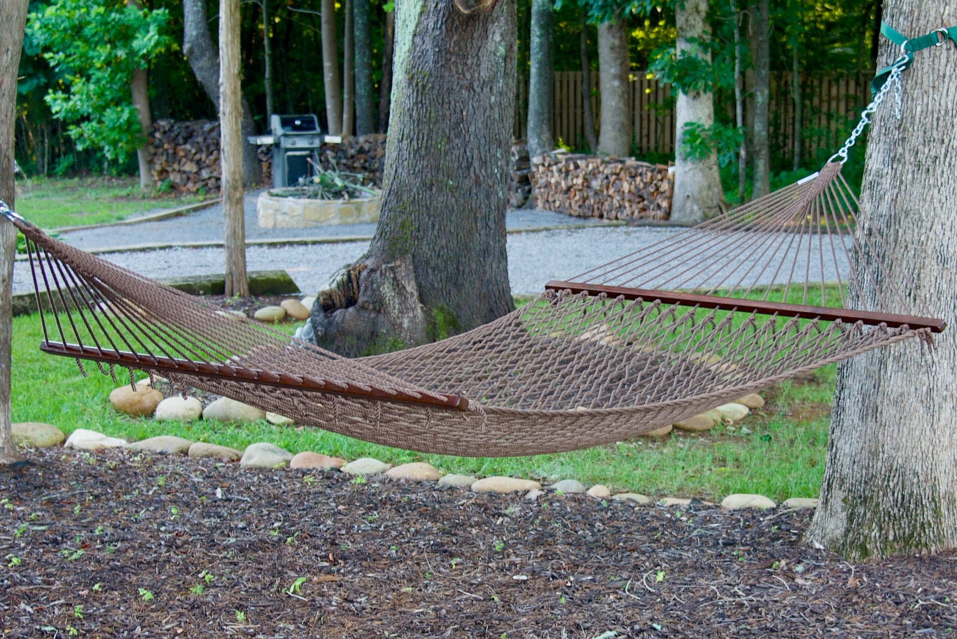 Mocha Island Escape hammock hung between two trees in a backyard with a fire pit and stacked firewood behind