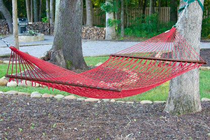 Red Island Escape hammock hung between two trees in a backyard with a fire pit and stacked firewood behind