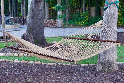 Tan Island Escape hammock hung between two trees in a backyard with a fire pit and stacked firewood behind