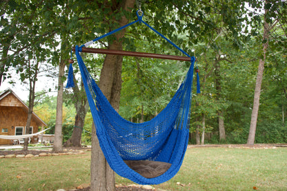 Dark blue Island Haven hammock chair with a brown seat pillow, hanging from a tree in a wooded backyard with a timber cabin behind