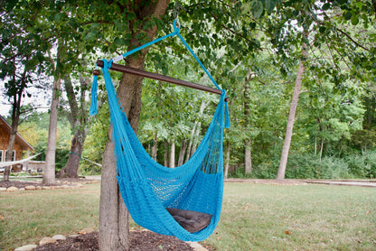Light blue Island Haven hammock chair with a brown seat pillow, hanging from a tree in a wooded backyard with a timber cabin behind