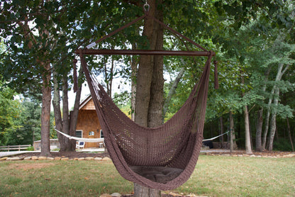 Mocha Island Haven hammock chair with a brown seat pillow, hanging from a tree in a wooded backyard with a timber cabin behind