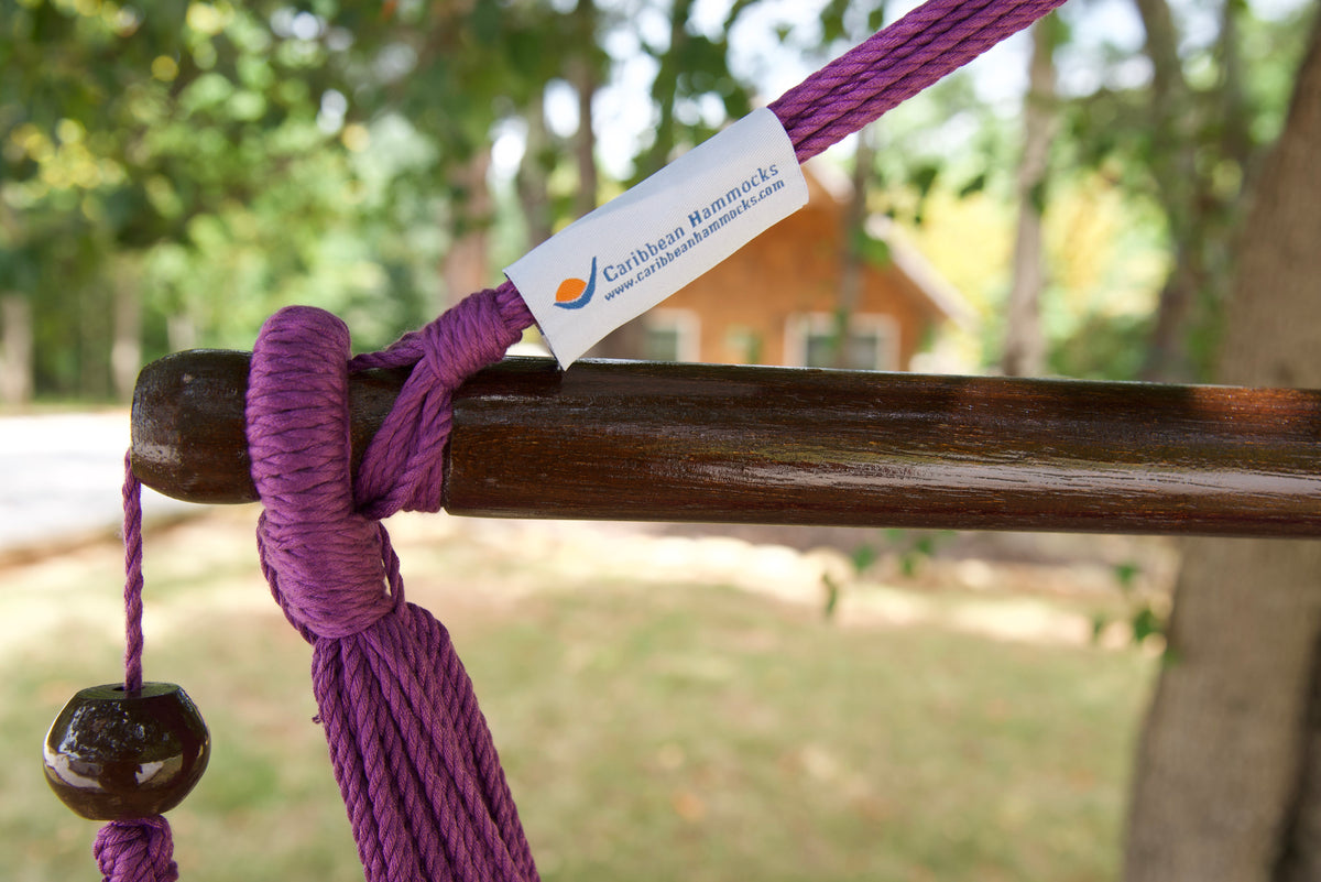 The dark varnished hardwood spreader bar of a purple Island Haven hammock chair, with end cords looped over it and a wooden bead hanging below