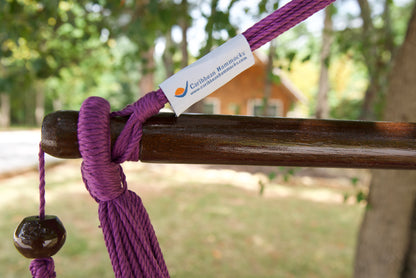 The dark varnished hardwood spreader bar of a purple Island Haven hammock chair, with end cords looped over it and a wooden bead hanging below