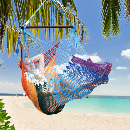 Woman resting in a rainbow-striped Island Haven hammock chair hung from a palm tree above a white sand beach