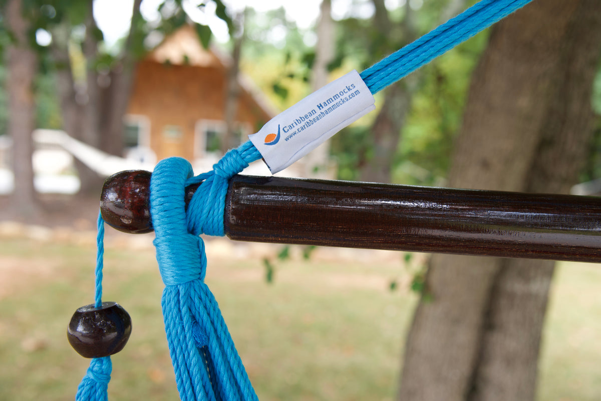 The dark varnished hardwood spreader bar of a rainbow-striped Island Haven hammock chair, with blue end cords looped over it and a wooden bead hanging below