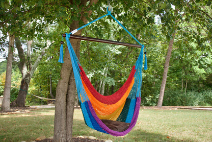 Rainbow-striped Island Haven hammock chair with a brown seat pillow, hanging from a tree in a wooded backyard with a timber cabin behind