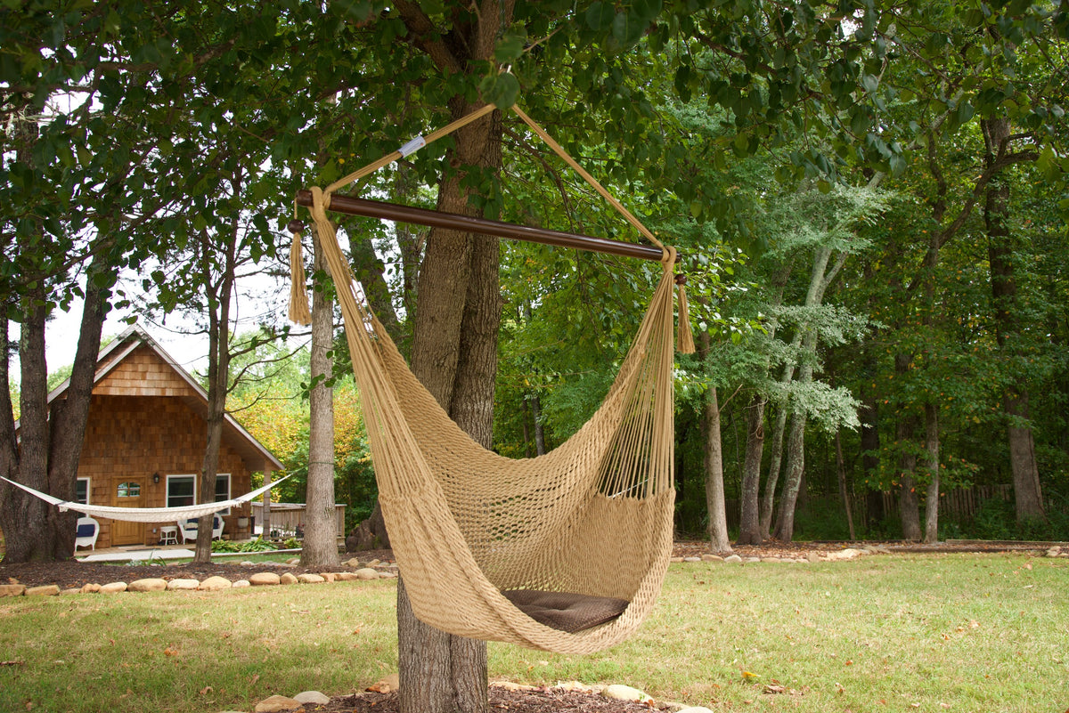 Tan Island Haven hammock chair with a brown seat pillow, hanging from a tree in a wooded backyard with a timber cabin behind