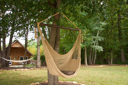 Tan Island Haven hammock chair with a brown seat pillow, hanging from a tree in a wooded backyard with a timber cabin behind