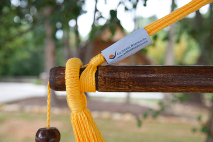 The dark varnished hardwood spreader bar of a yellow Island Haven hammock chair, with end cords looped over it and a wooden bead hanging below