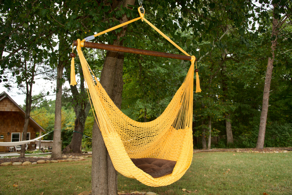 Yellow Island Haven hammock chair with a brown seat pillow, hanging from a tree in a wooded backyard with a timber cabin behind