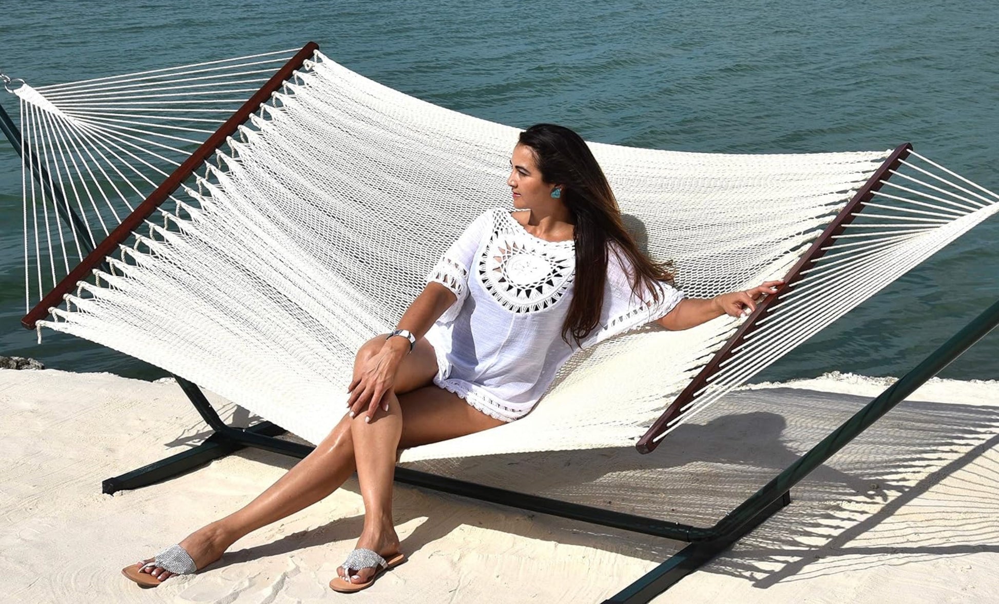 Woman in white sitting on a cream Island Oasis hammock on a black steel hammock stand beside the ocean