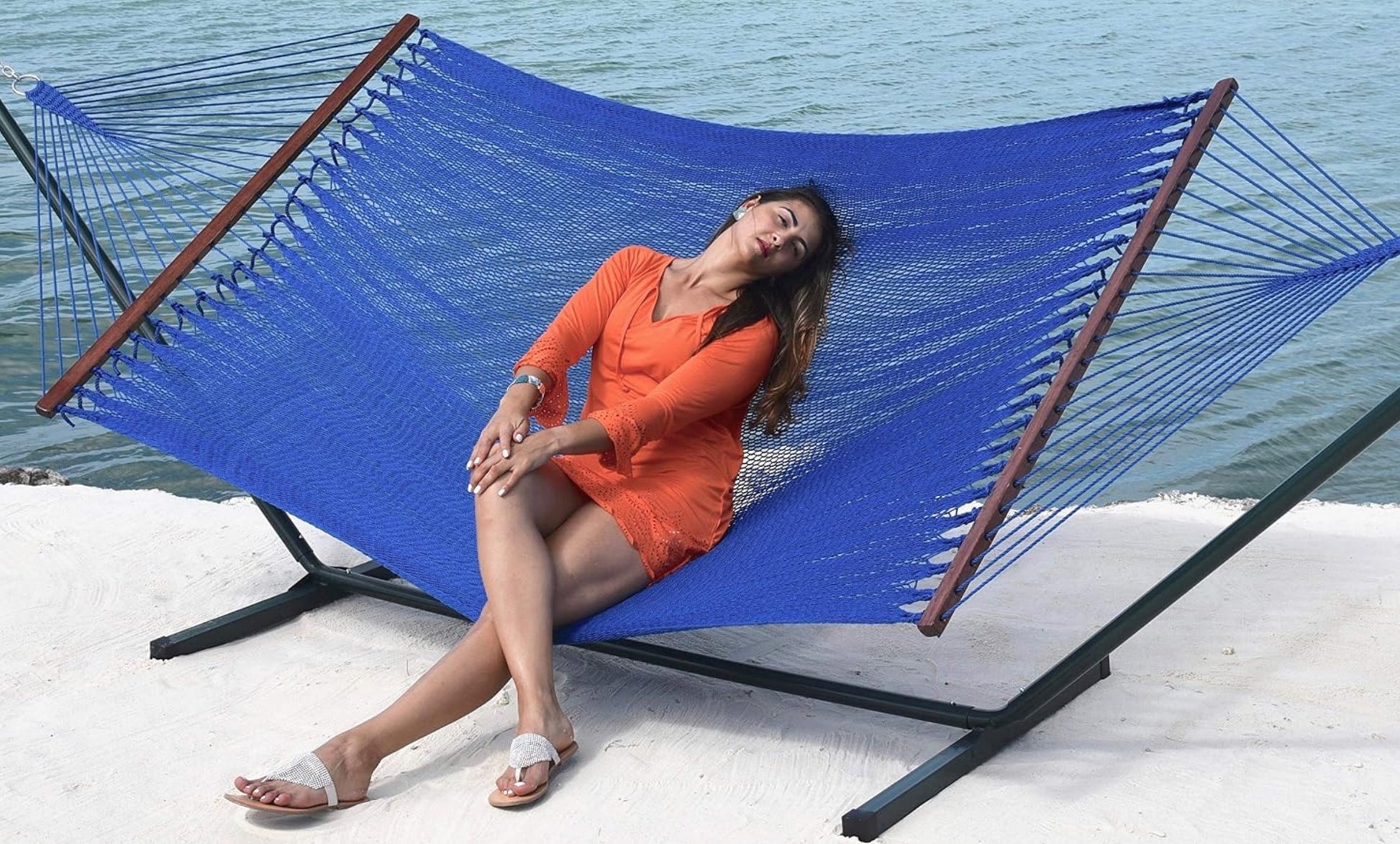 Woman in an orange dress reclining in a dark blue Island Oasis hammock on a black steel hammock stand beside the ocean
