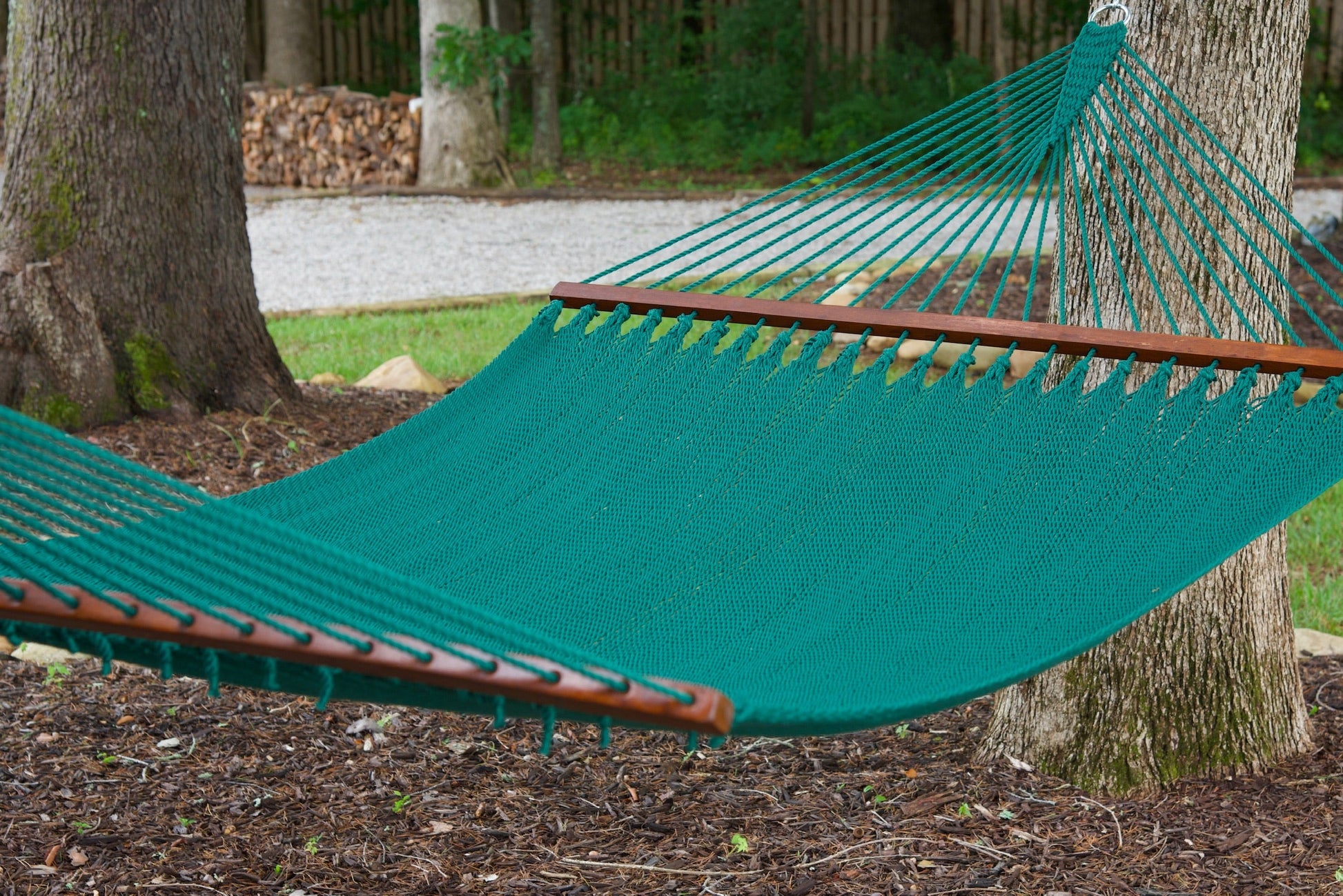 Green Island Oasis hammock hung between two trees above a mulched bed, woodpile and fence behind