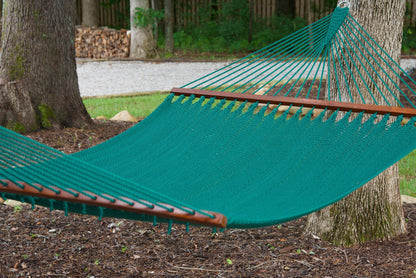Green Island Oasis hammock hung between two trees above a mulched bed, woodpile and fence behind