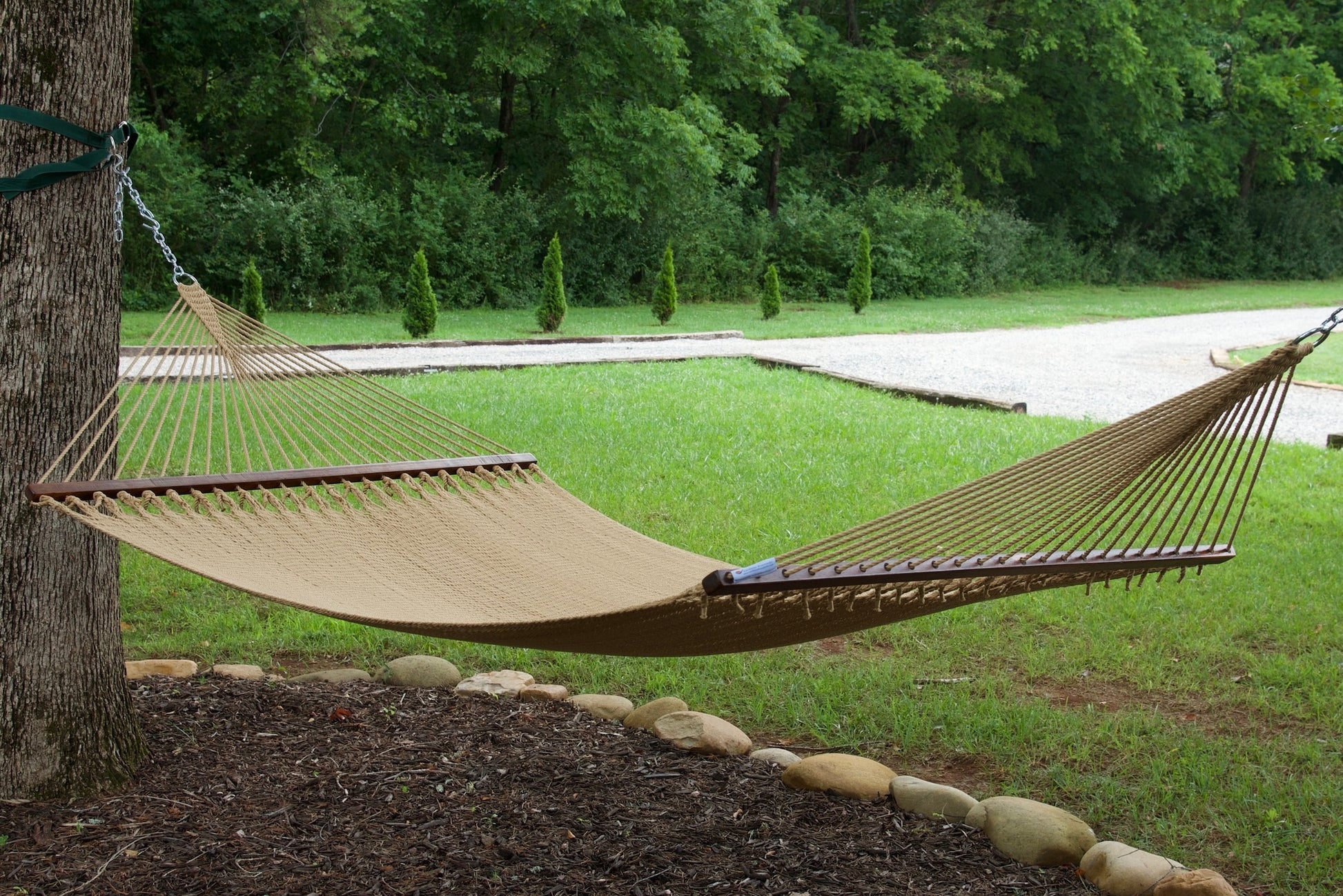 Tan Island Oasis hammock hung between a tree and a post in a green backyard with a gravel path behind