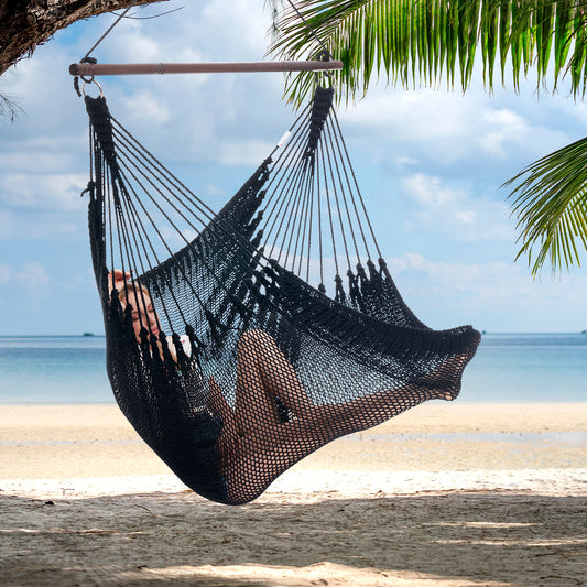 Person reclining in a black Jumbo Caribbean Hammock Chair hung from a palm tree above a white sand beach