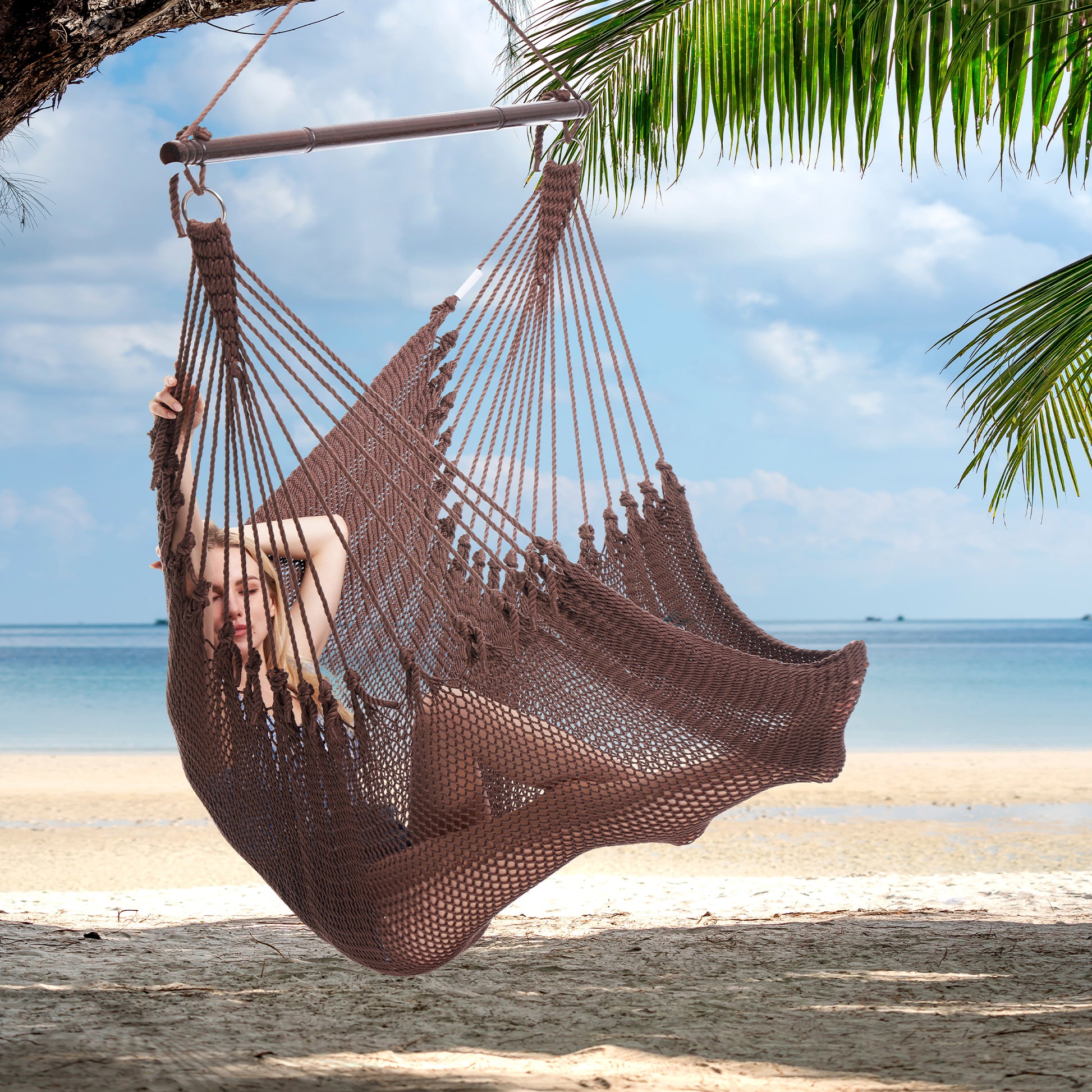Person reclining in a mocha Jumbo Caribbean Hammock Chair hung from a palm tree above a white sand beach