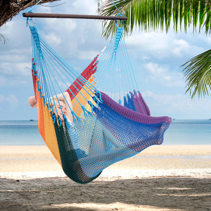Person reclining in a rainbow Jumbo Caribbean Hammock Chair hung from a palm tree above a white sand beach