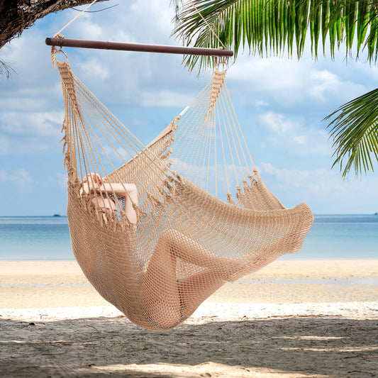 Person reclining in a tan Jumbo Caribbean Hammock Chair hung from a palm tree above a white sand beach