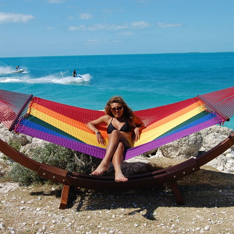 Woman seated in the Rainbow Jumbo Caribbean Hammock on the Wood Arc Hammock Stand beside the ocean