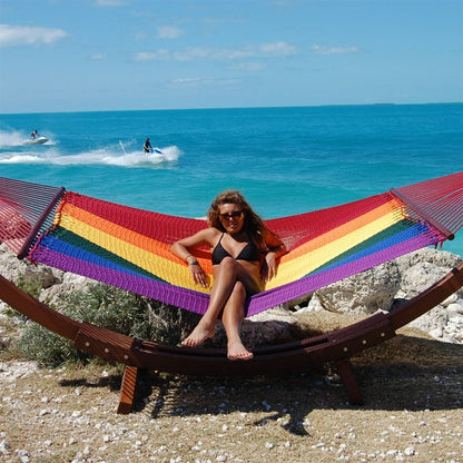 Woman seated in the Rainbow Jumbo Caribbean Hammock on the Wood Arc Hammock Stand beside the ocean