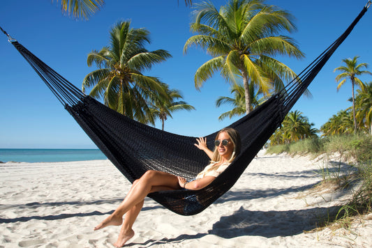 Black Jumbo Mayan Hammock strung between palm trees on a white-sand beach, a woman sitting in it with her feet on the sand