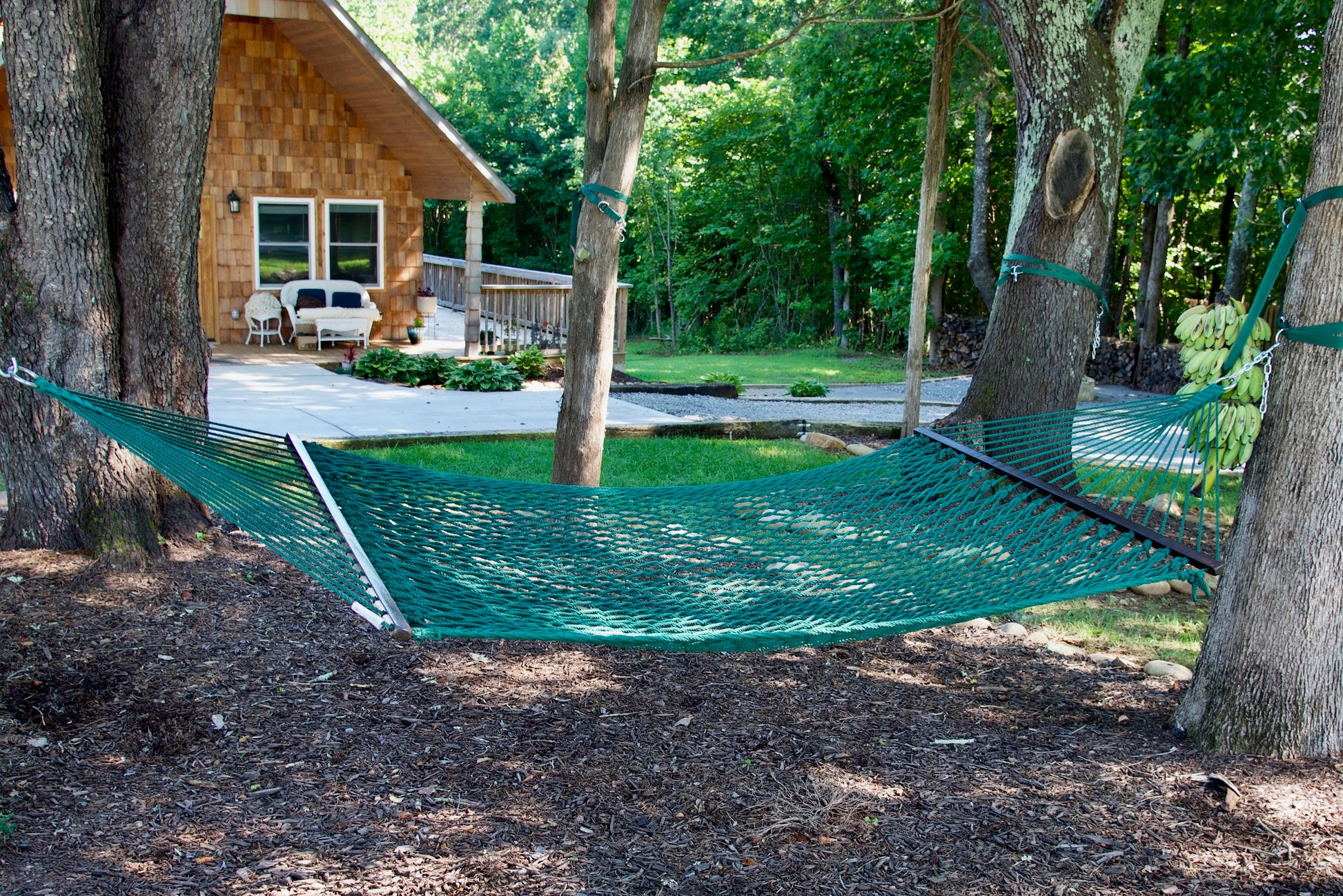 Green Jumbo Rope Hammock hanging between two trees in a landscaped backyard.