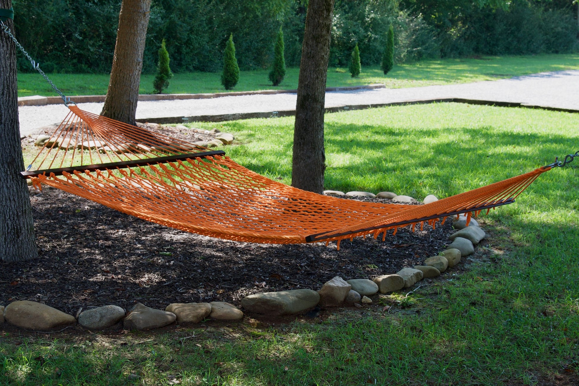 Orange Jumbo Rope Hammock hanging between two trees in a landscaped backyard.