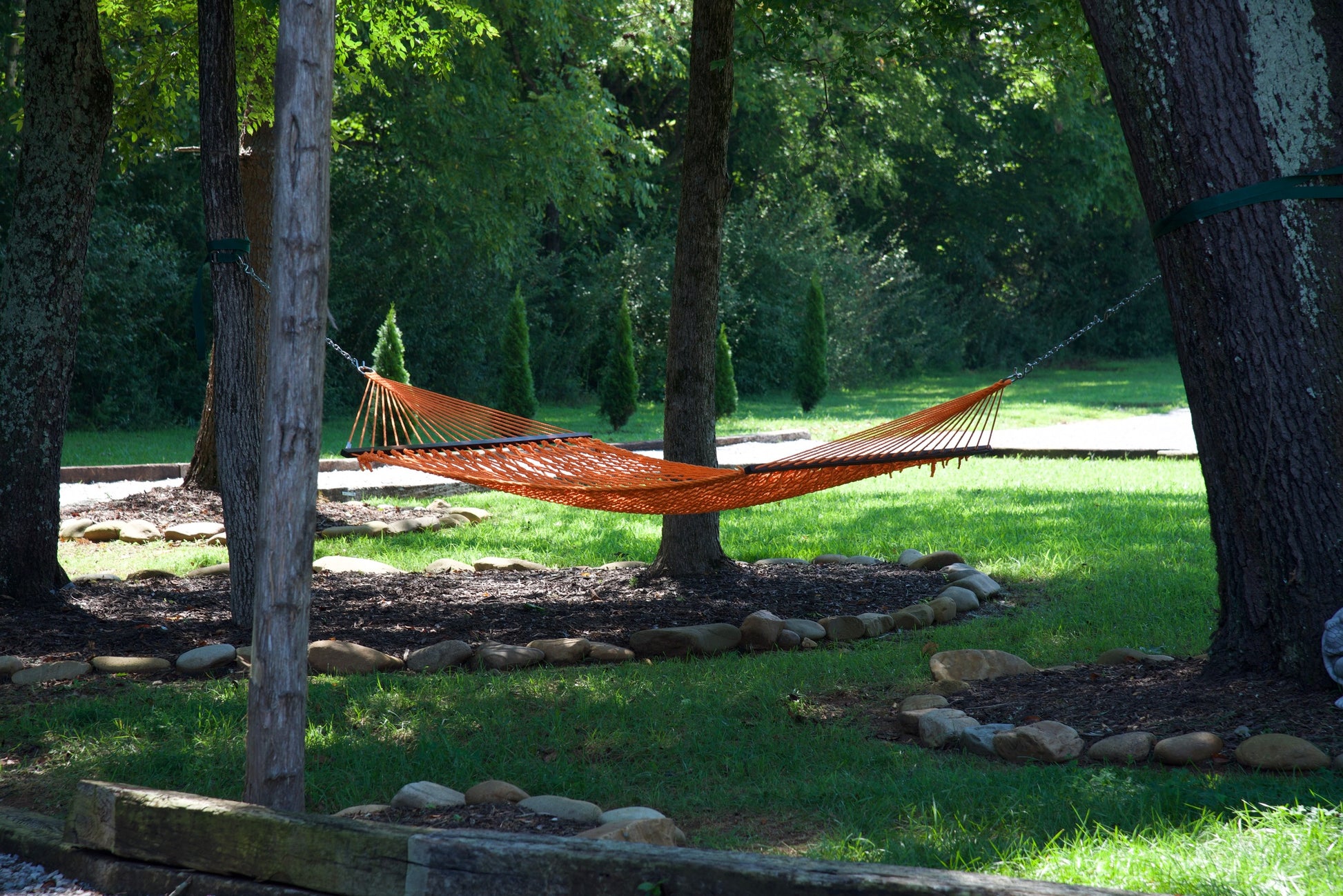 Orange Jumbo Rope Hammock hanging between two trees in a landscaped backyard.