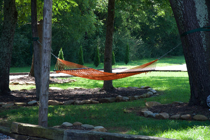 Orange Jumbo Rope Hammock hanging between two trees in a landscaped backyard.