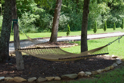 Tan Jumbo Rope Hammock hanging between two trees in a landscaped backyard.