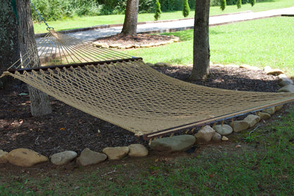 Tan Jumbo Rope Hammock hanging between two trees in a landscaped backyard.