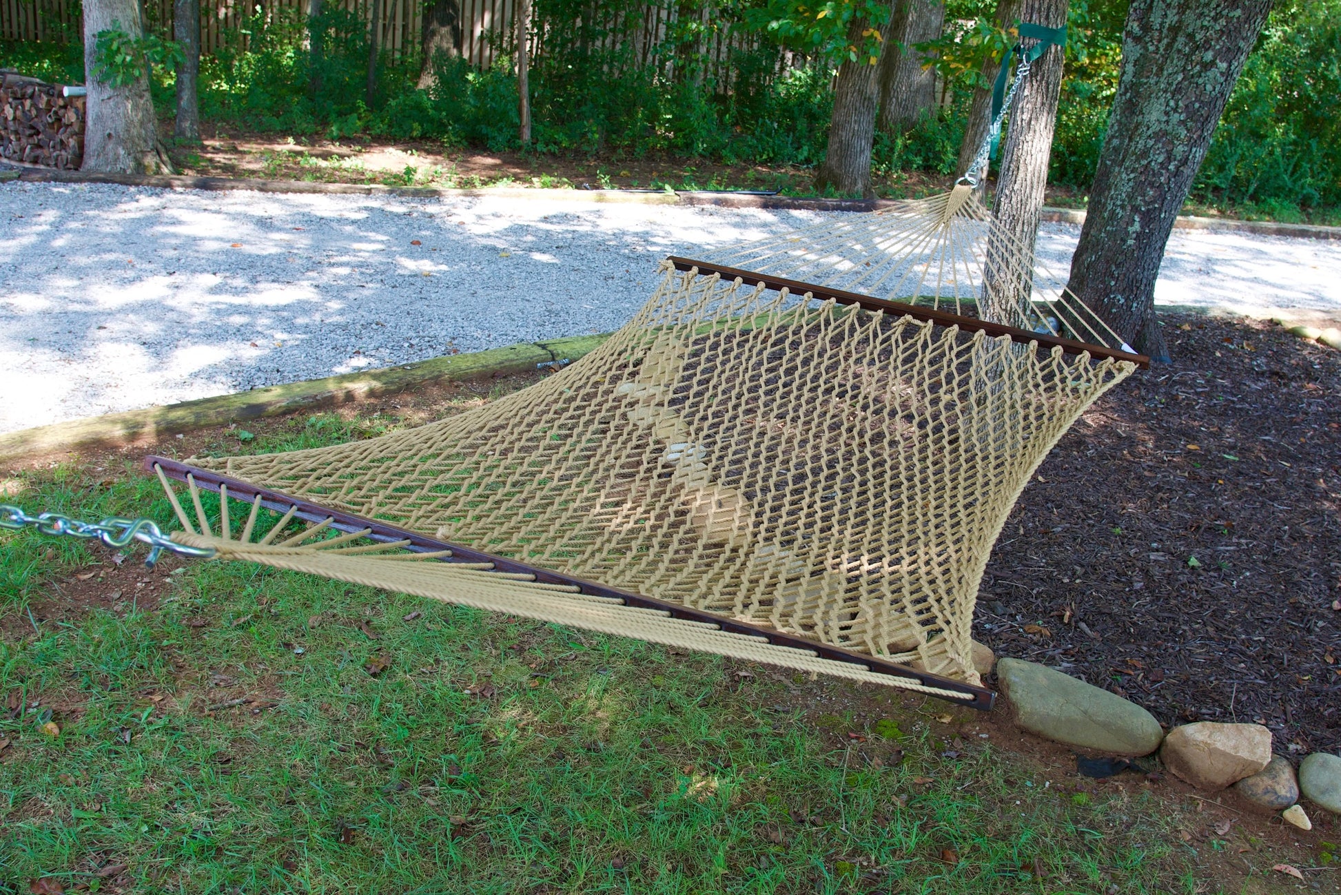 Tan Jumbo Rope Hammock hanging between two trees in a landscaped backyard.