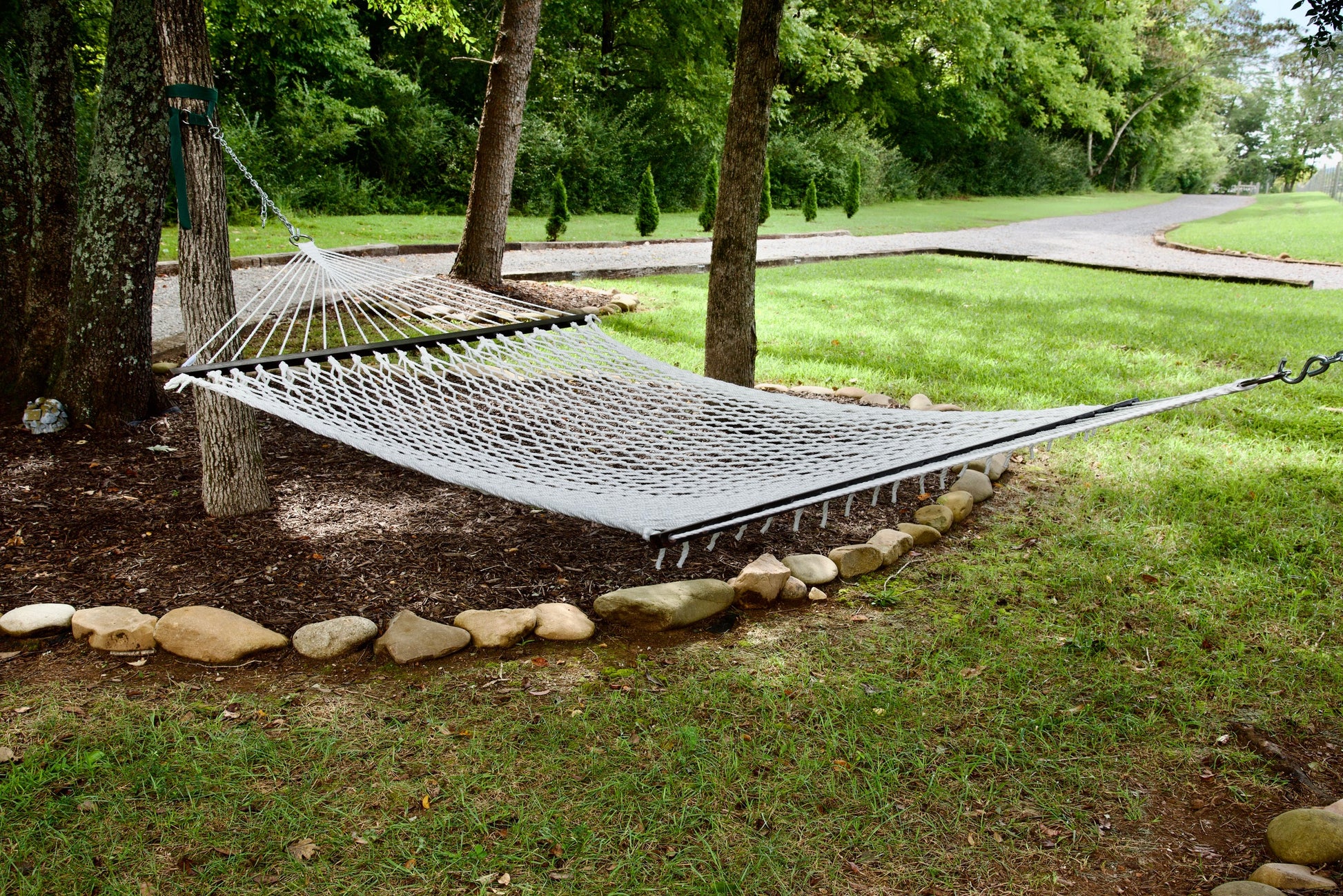 White Jumbo Rope Hammock hanging between two trees in a landscaped backyard.
