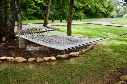 White Jumbo Rope Hammock hanging between two trees in a landscaped backyard.