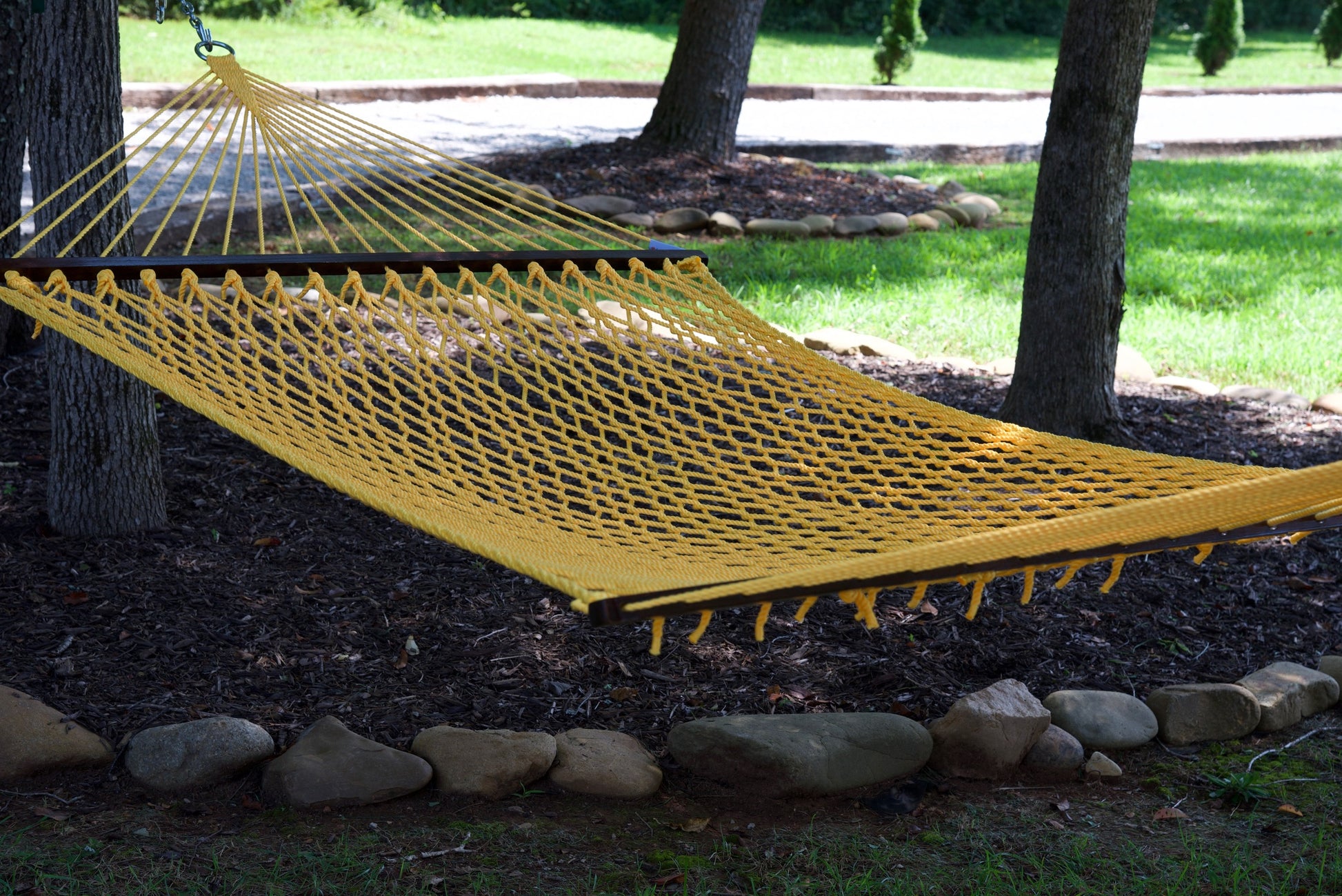 Yellow Jumbo Rope Hammock hanging between two trees in a landscaped backyard.