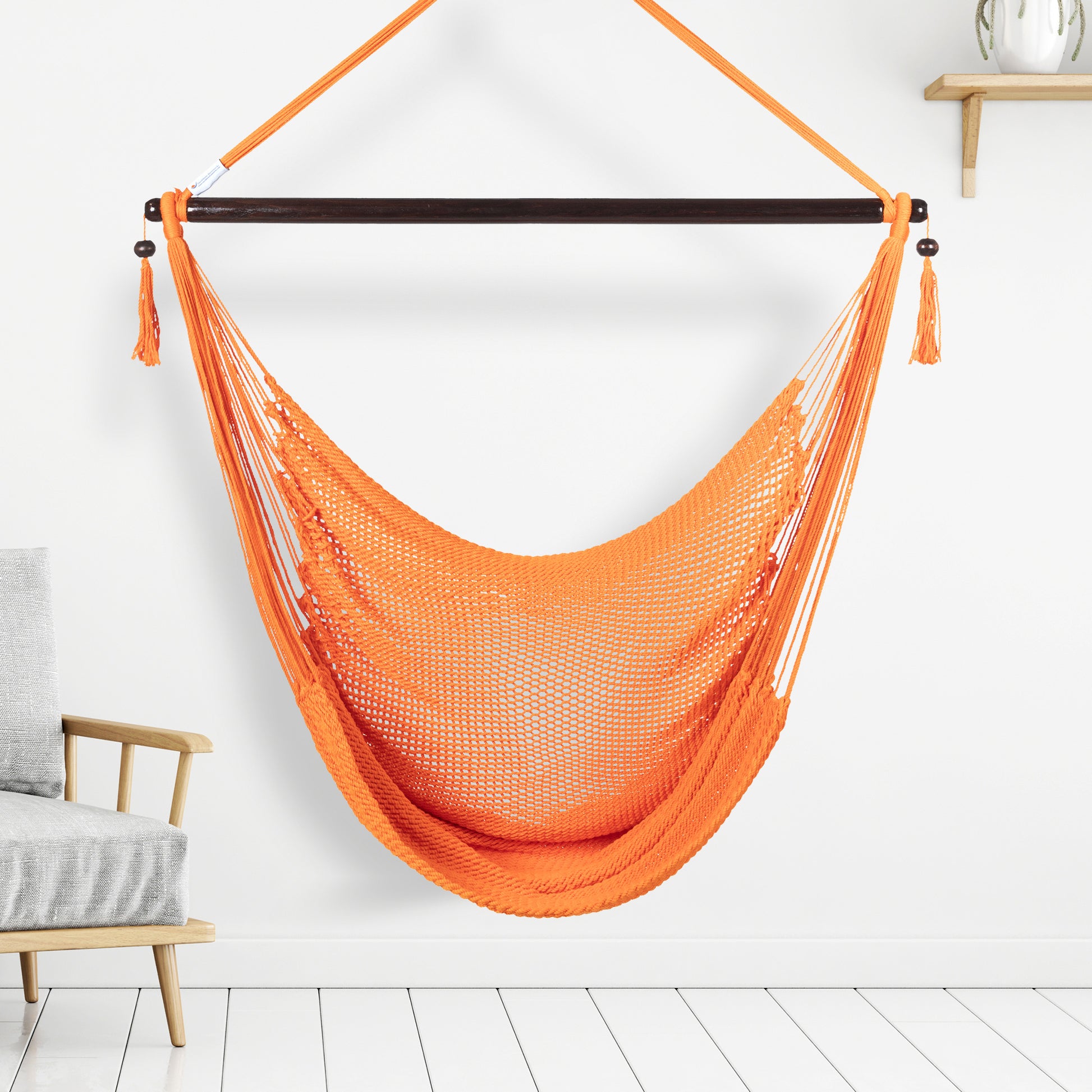 Orange Large Caribbean Hammock Chair hanging indoors beside a pale wood armchair against a white wall