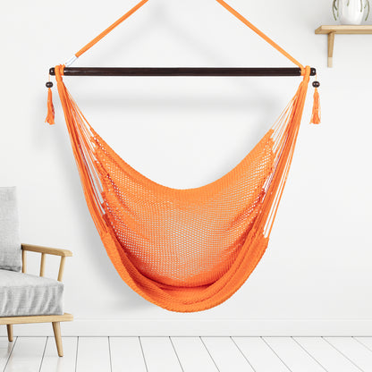 Orange Large Caribbean Hammock Chair hanging indoors beside a pale wood armchair against a white wall