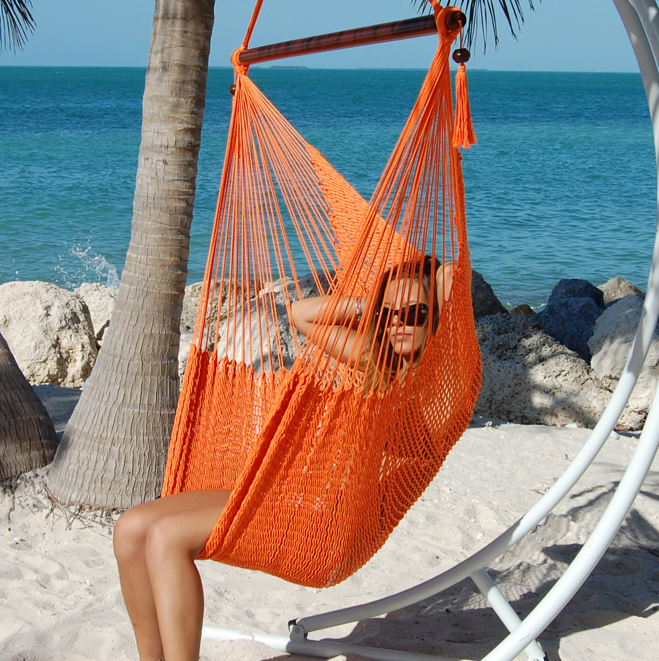 Woman seated in an orange Large Caribbean Hammock Chair on a white arc stand on a sandy beach