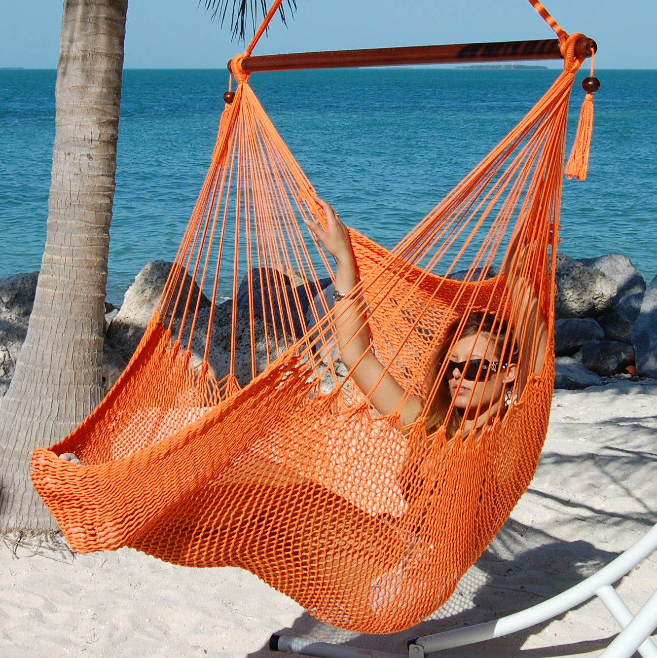 Woman reclining in an orange Large Caribbean Hammock Chair on a white arc stand beside turquoise water