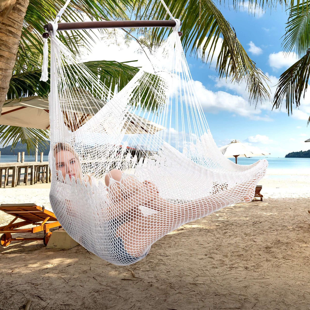 Woman lying back in a white Large Caribbean Hammock Chair on a tropical beach with loungers and palm fronds