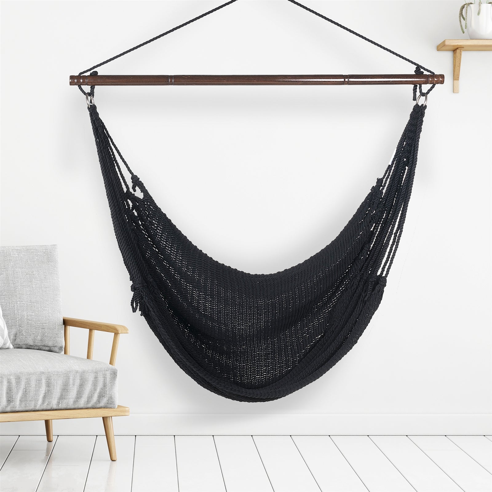 Black Jumbo Caribbean Hammock Chair hanging indoors beside a pale wood armchair against a white wall