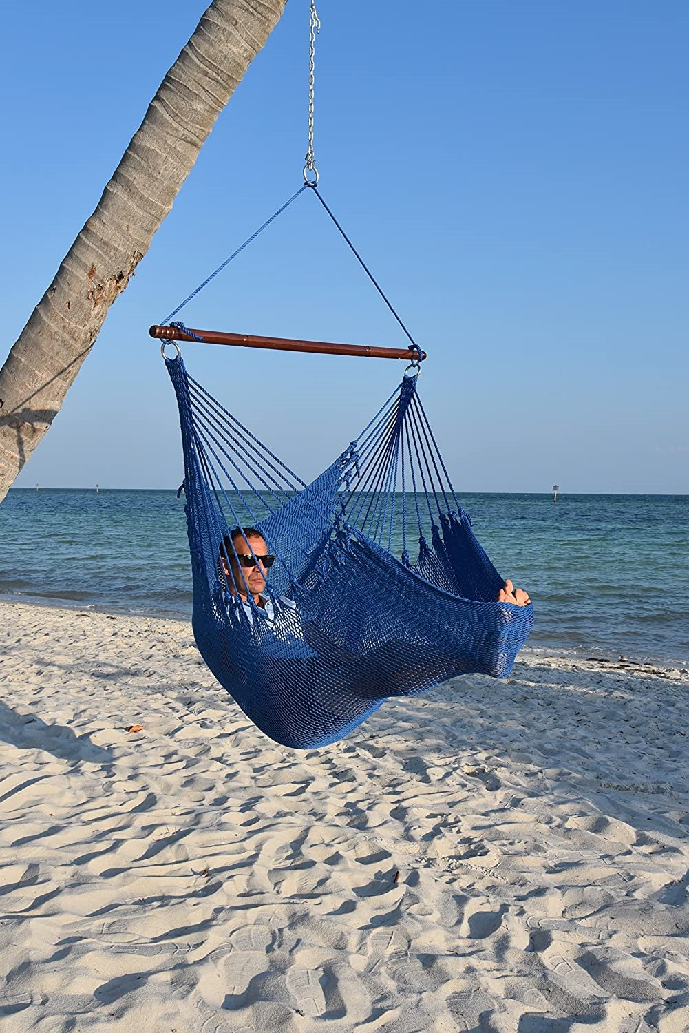 Man relaxing in a dark blue Jumbo Caribbean Hammock Chair hung from a leaning palm tree on a beach