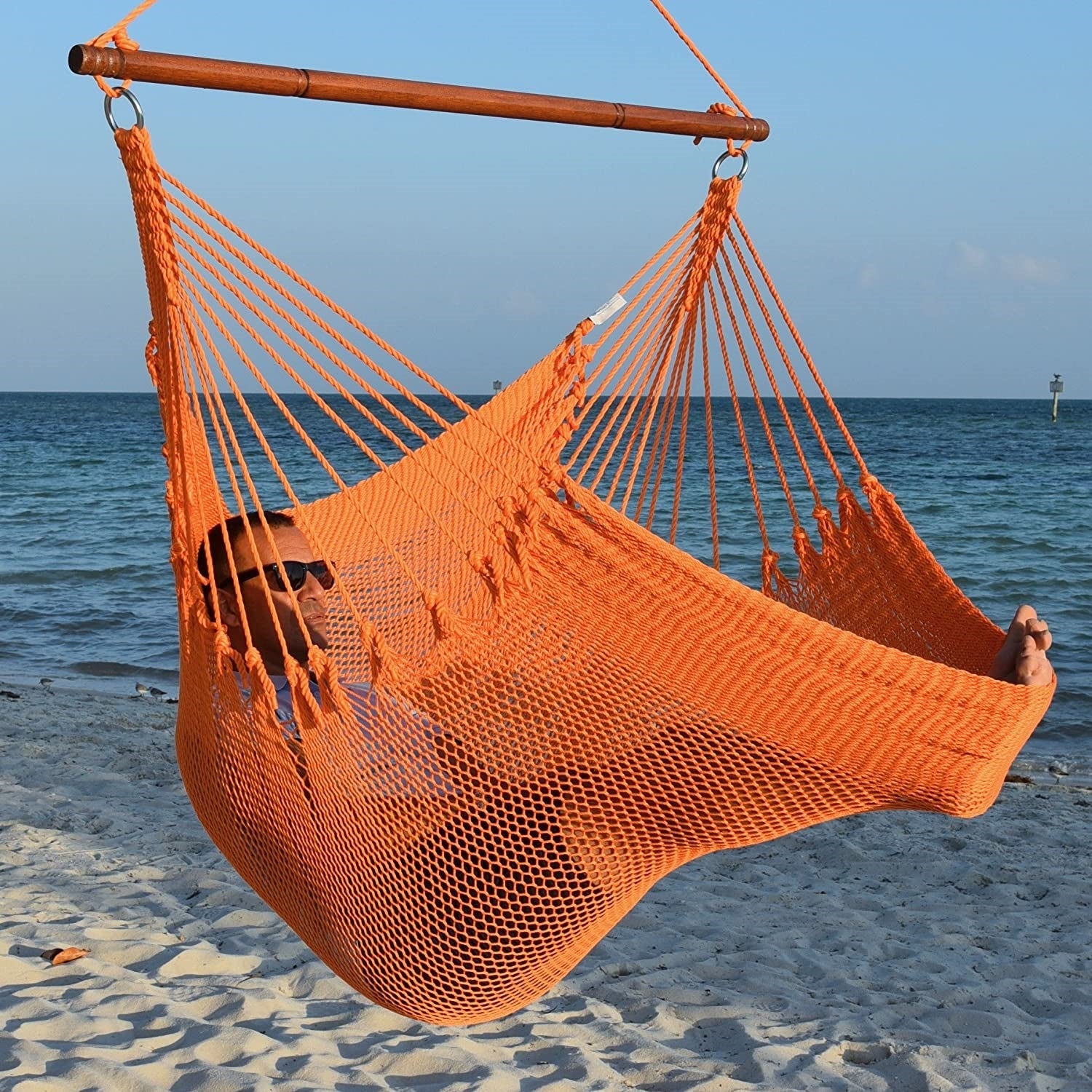 Man in sunglasses stretched out in an orange Jumbo Caribbean Hammock Chair with calm ocean behind