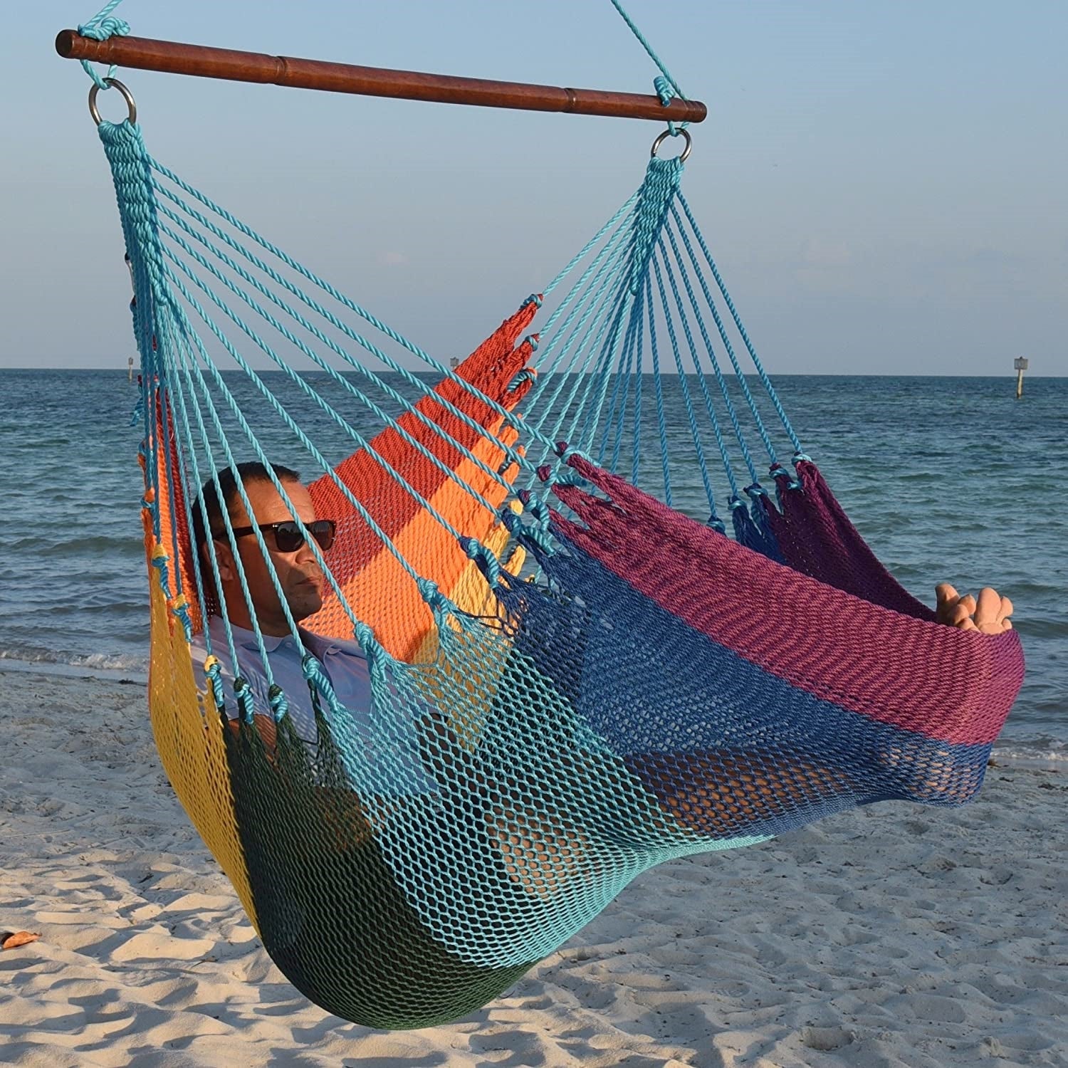 Man in sunglasses stretched out in a rainbow Jumbo Caribbean Hammock Chair with calm ocean behind